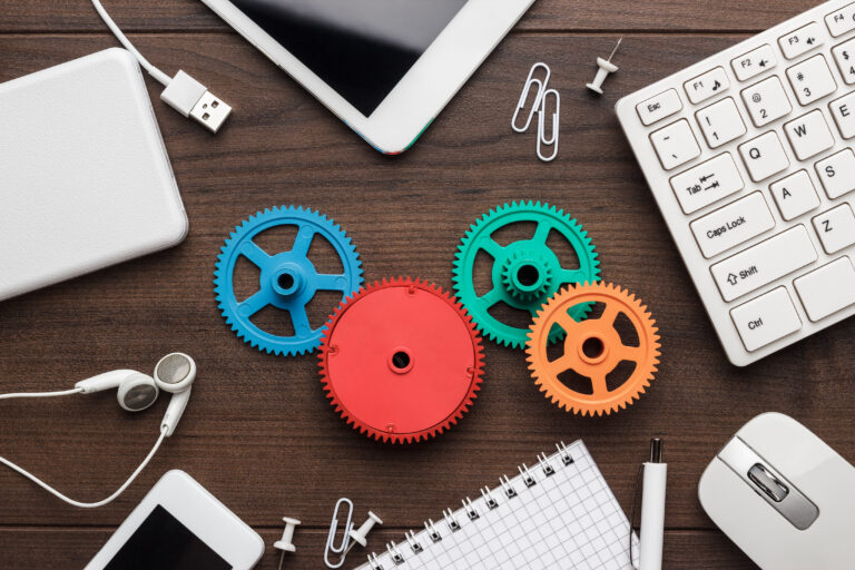 A set of plastic gears placed on a brown desk beside a keyboard, mouse, tablet, headset, and notebook.