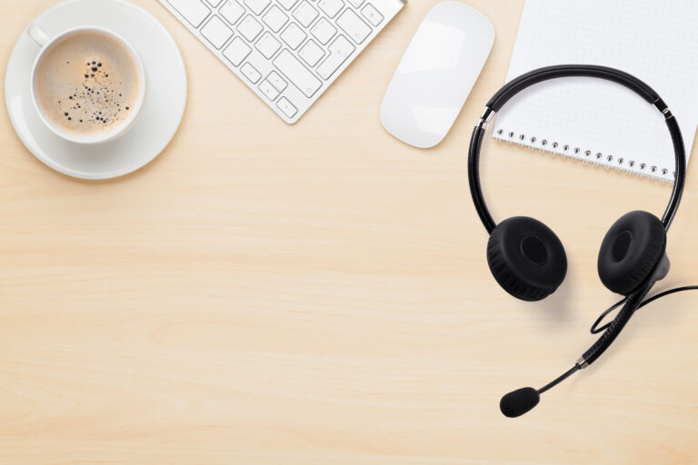 A wooden office table with coffee, mouse, keyboard, headset, and notes on top of it.