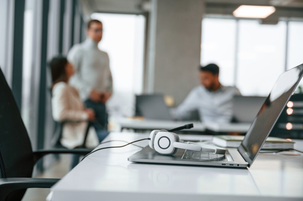 A closeup shot of headphone lying on top of a laptop with employees working in the background.