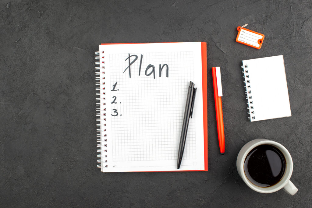 Overhead view of a notebook with a plan and numbers written on it beside a pen, smaller notebook and a coffee mug.