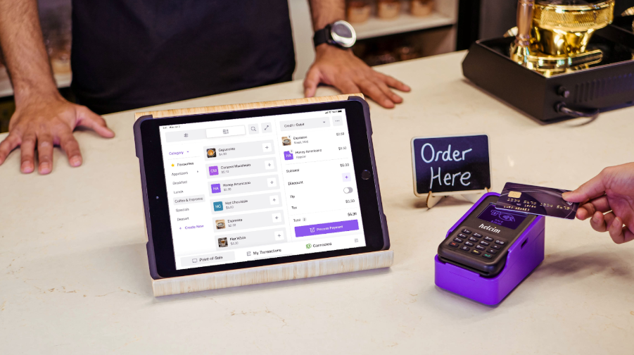 Customer tapping a credit card on a Helcim reader beside a tablet POS system at a coffee shop counter