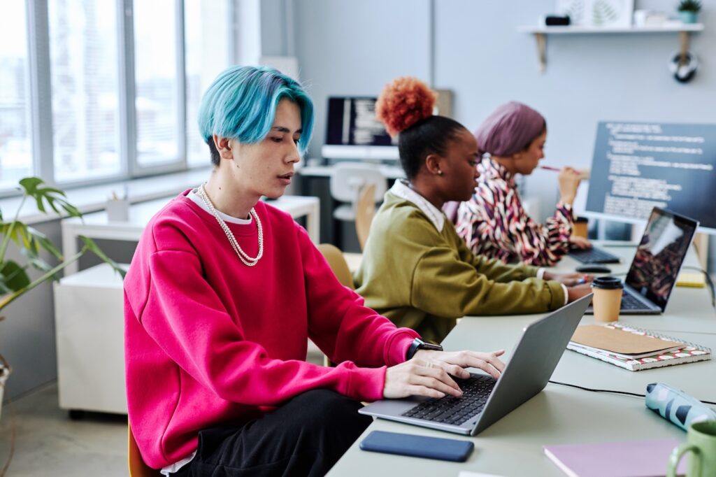 Diverse team of gen Z software developers working in office; focus on Asian young man with colored hair using computer in foreground.