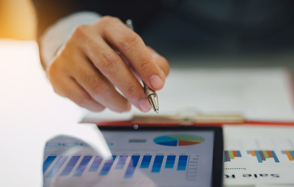 Close-up of a person's hand holding a pen over a tablet displaying financial growth charts and sales reports.