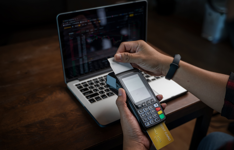 A person inserting a yellow credit card into a payment terminal next to a laptop displaying trading charts.