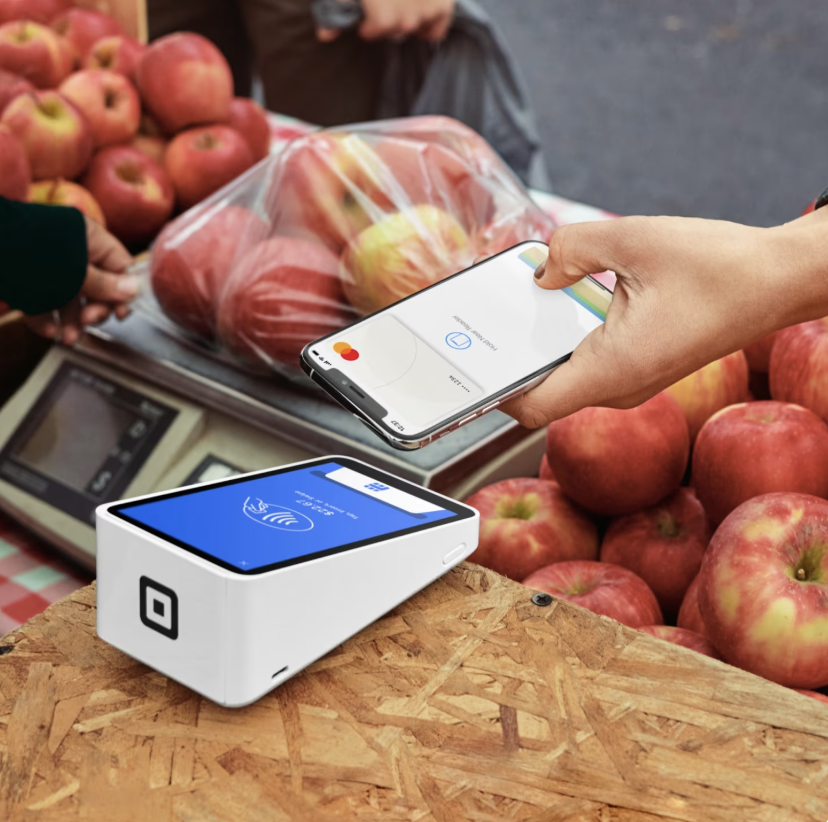 Customer using a smartphone to make a contactless payment at a market beside a Square card reader.