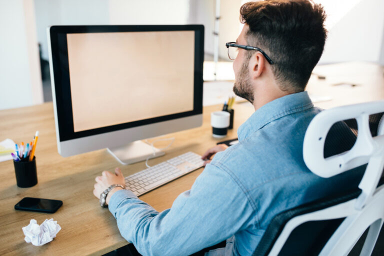 a man is working with a computer at his desktop in office. He wears blue shirt and looks busy. View from back.